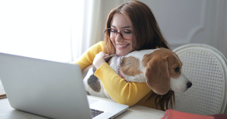 woman sitting at a table holding a dog while looking at a laptop