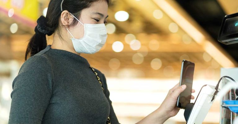image of a woman wearing a medical face mask holds her phone up to a credit card terminal to make a touchless payment